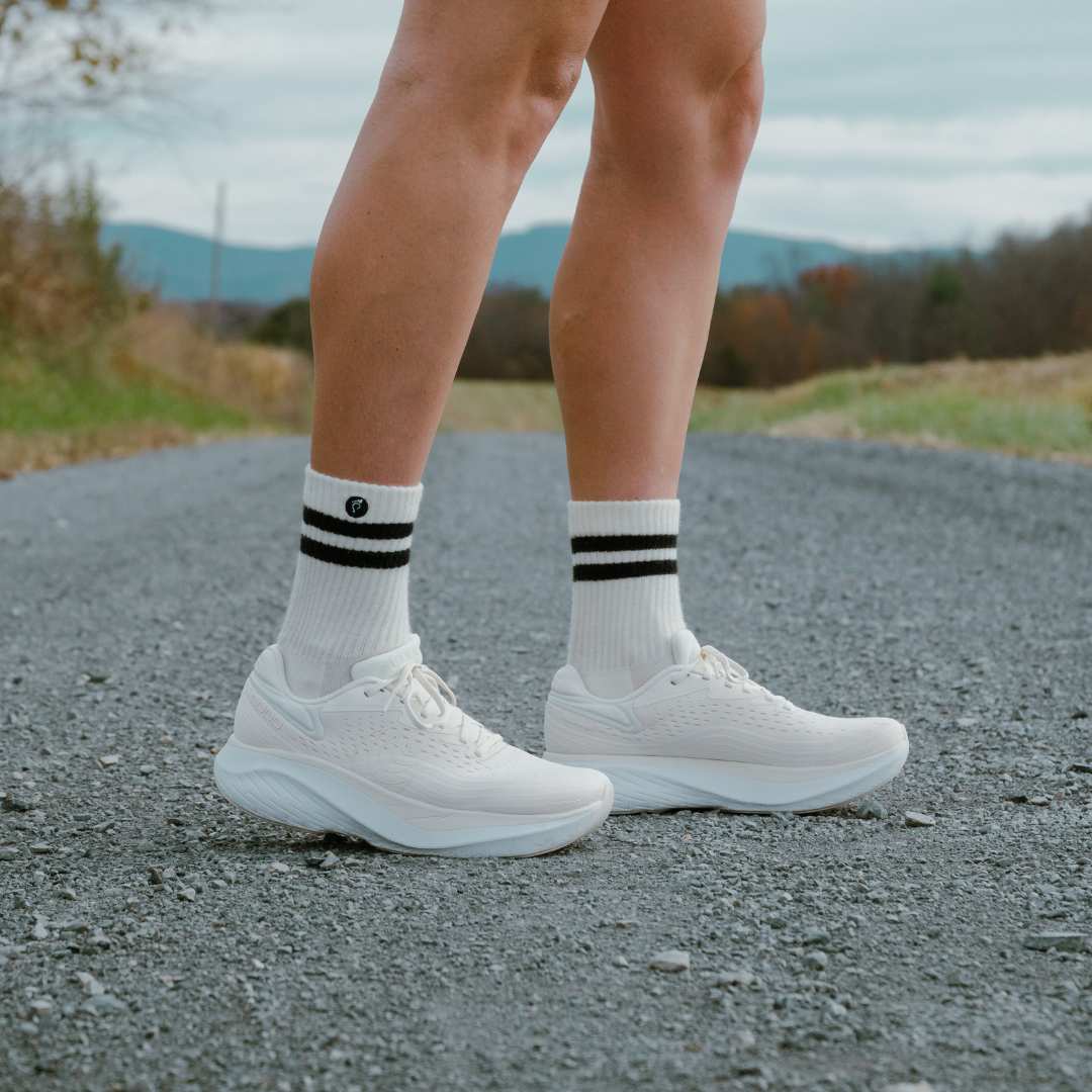 A person wearing white running shoes and striped socks stands on a gravel road.