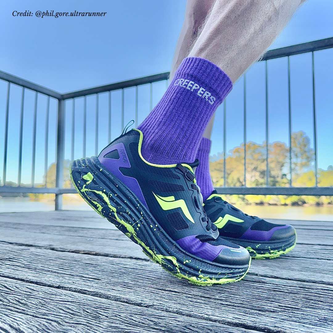 A person wearing purple socks and black and purple running shoes with neon green accents on a wooden deck.