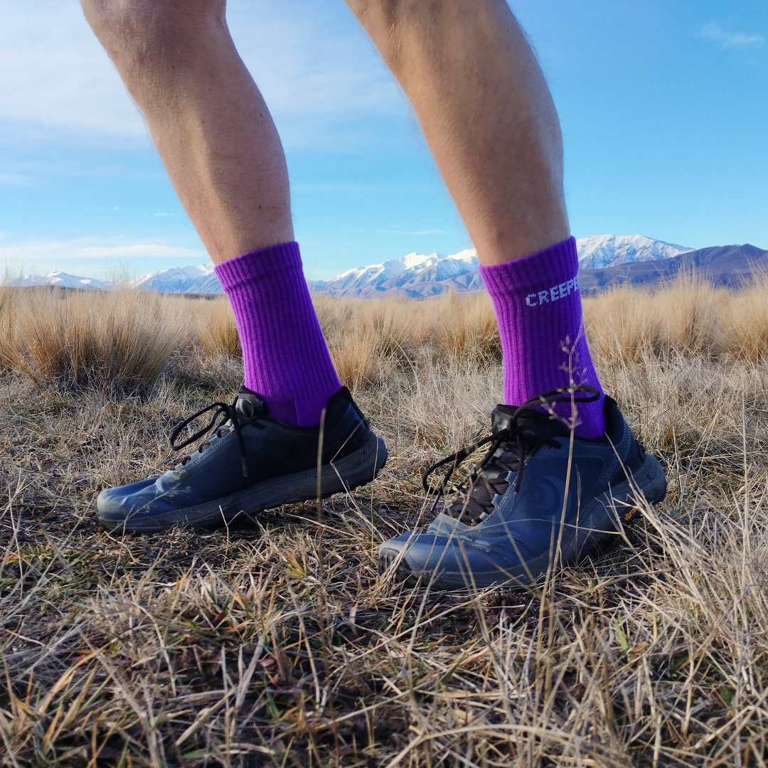 A person wearing light-colored trail shoes and green socks stands on a rocky, leaf-covered path.