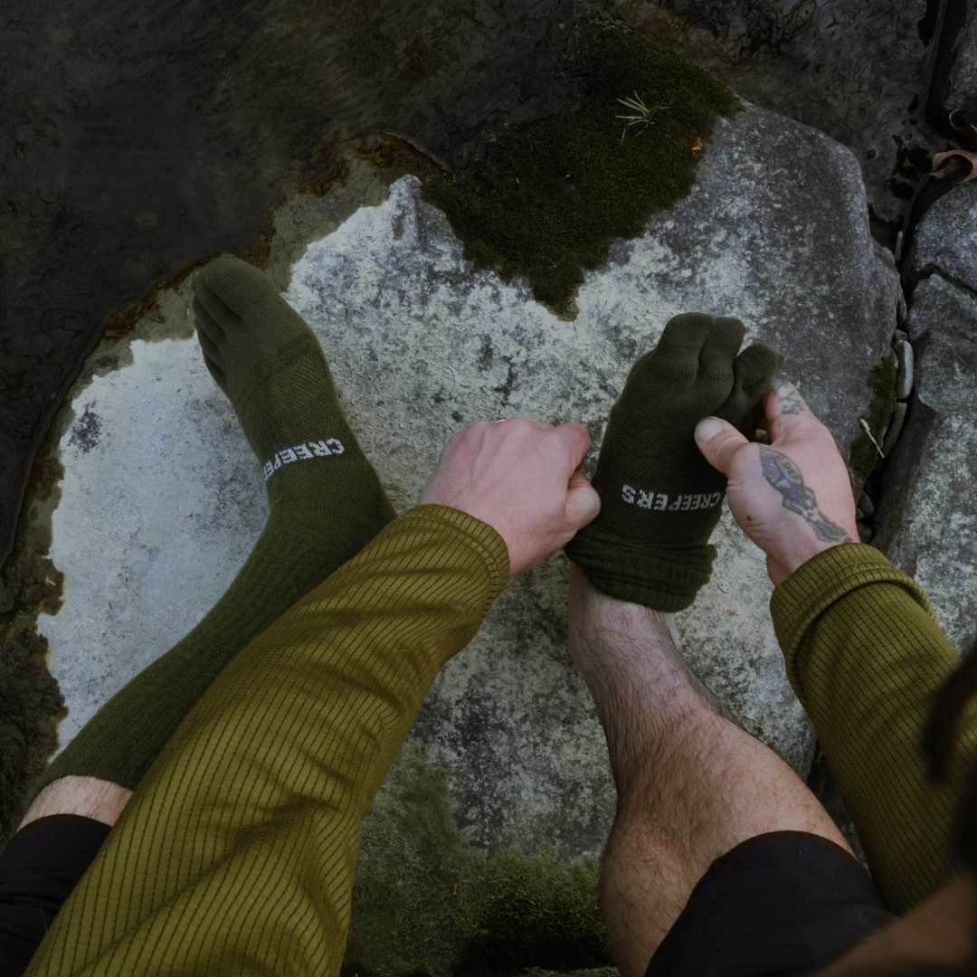 A person with a tattooed hand puts on a pair of green toe socks while sitting on a rock.