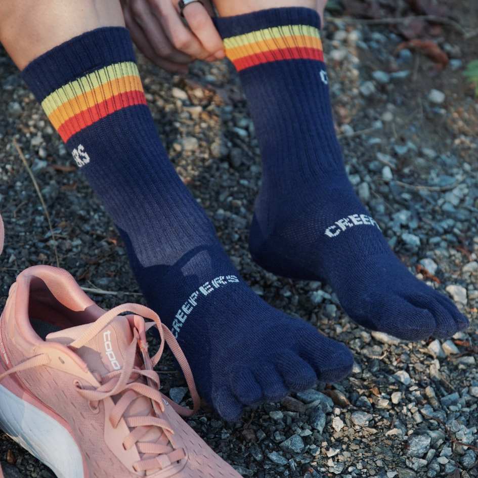 Person wearing toe socks with colorful stripes, next to pink sneakers on gravel.