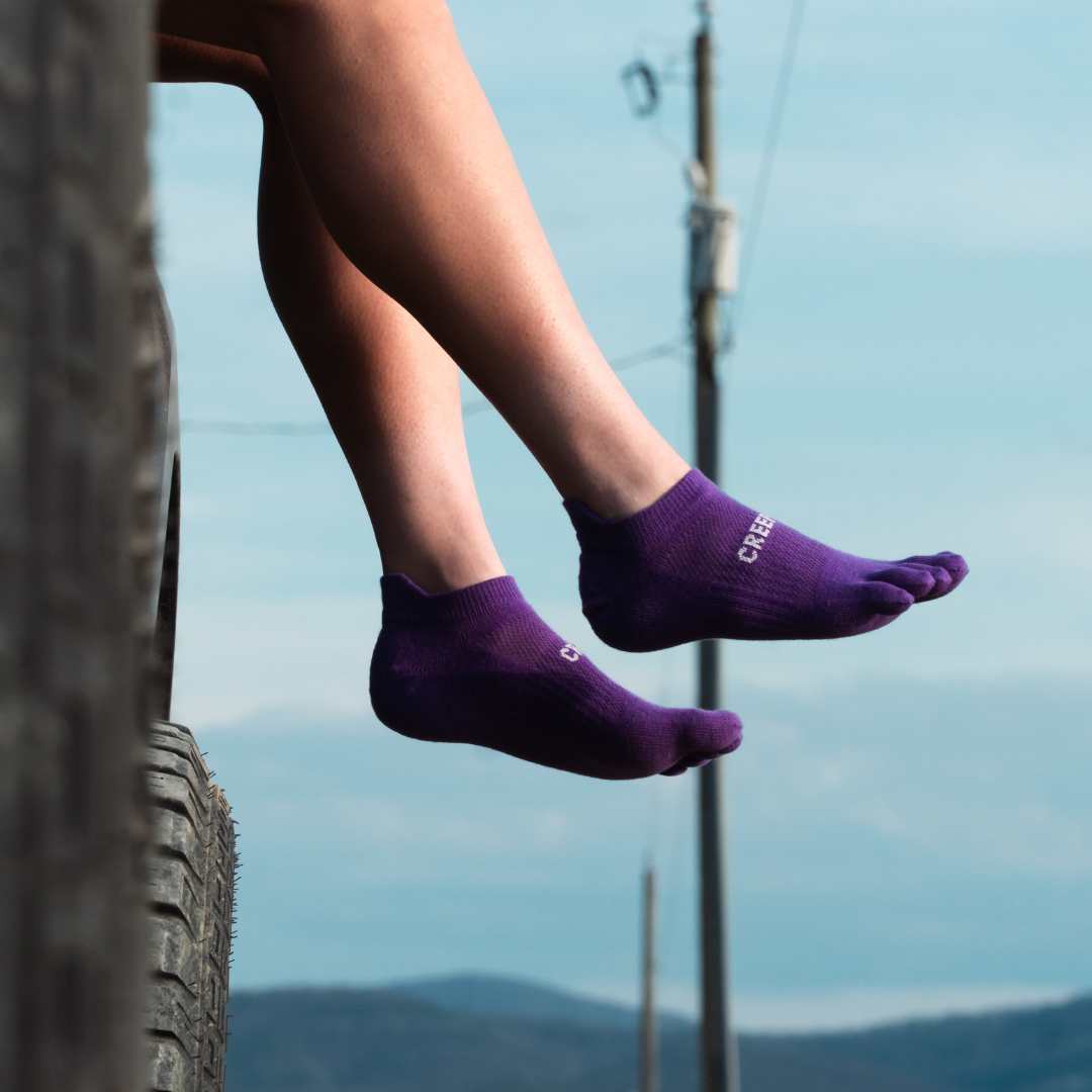 A person with a tattooed hand puts on a pair of green toe socks while sitting on a rock.