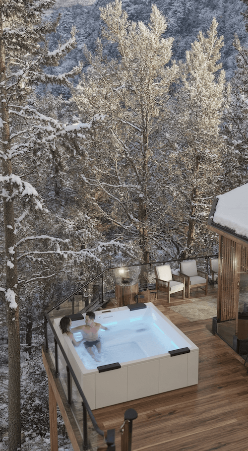 Two people in a hot tub on a snowy deck surrounded by trees.