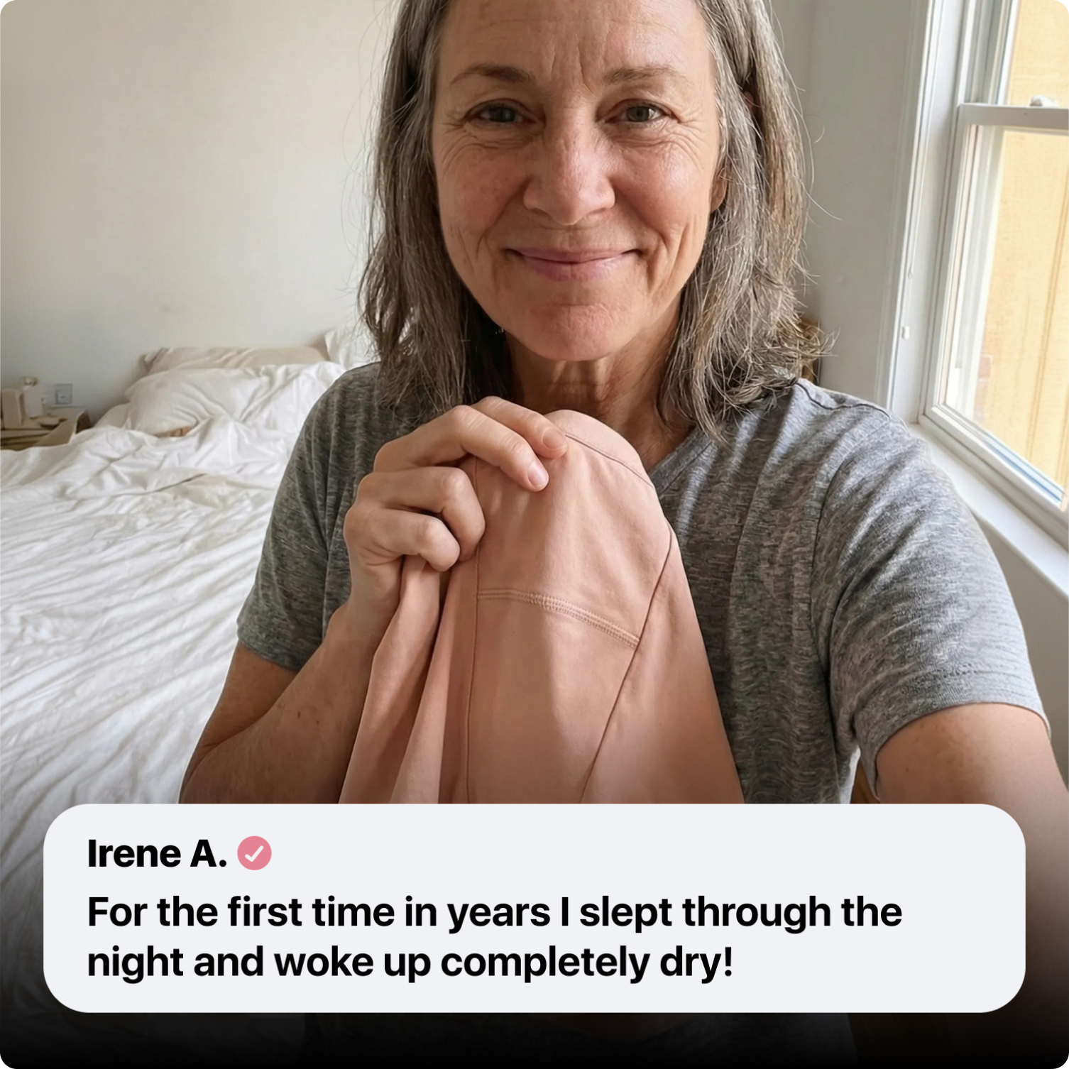 A smiling woman in a gray t-shirt holds up a piece of pink fabric in a bedroom.