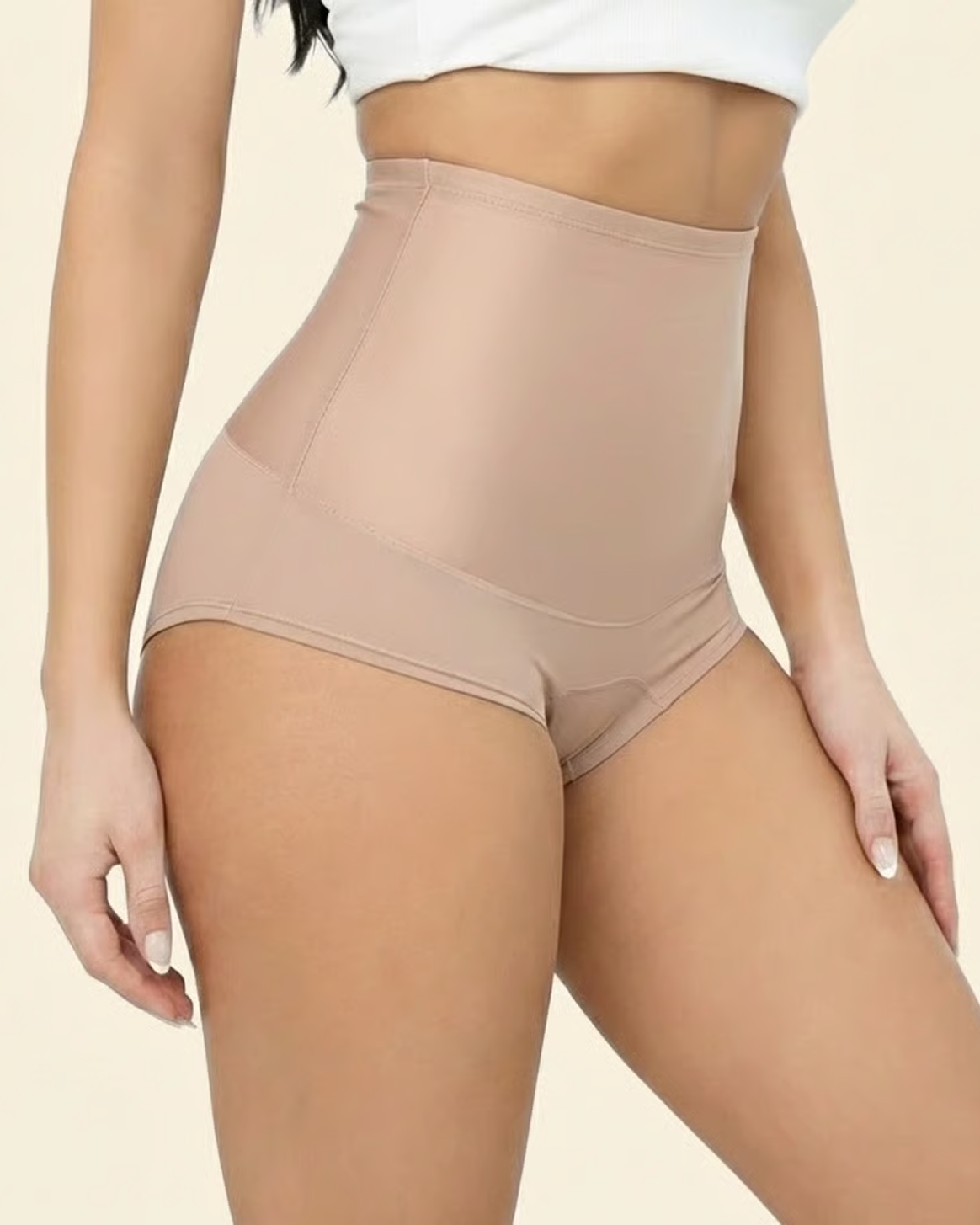 A woman models high-waisted, beige tummy control shapewear shorts against a solid background.