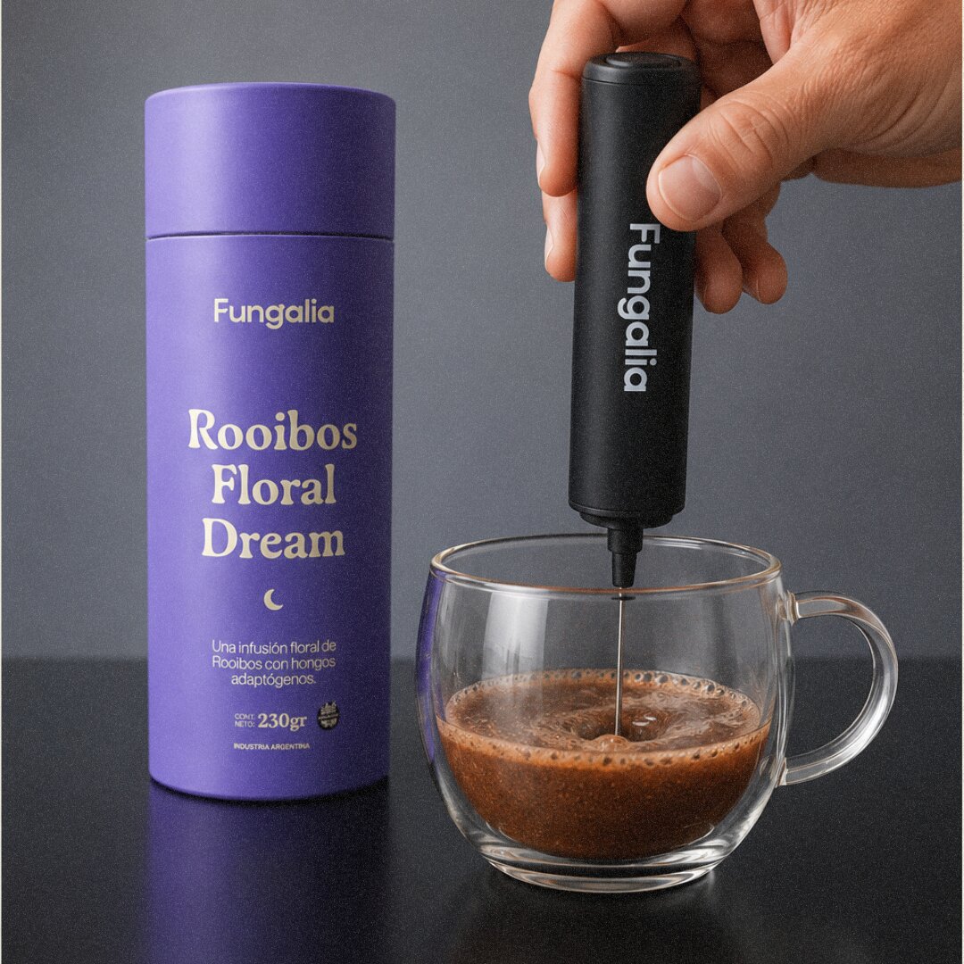 A hand uses a handheld frother to mix a drink in a glass mug next to a purple container.