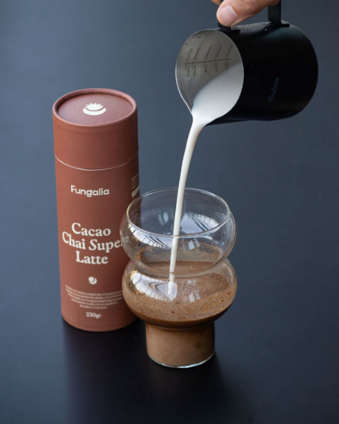 Milk being poured into a glass with cacao chai latte container nearby.