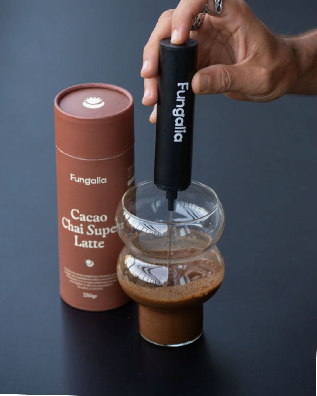 Hand using a frother in a glass of cacao chai latte next to a product container.