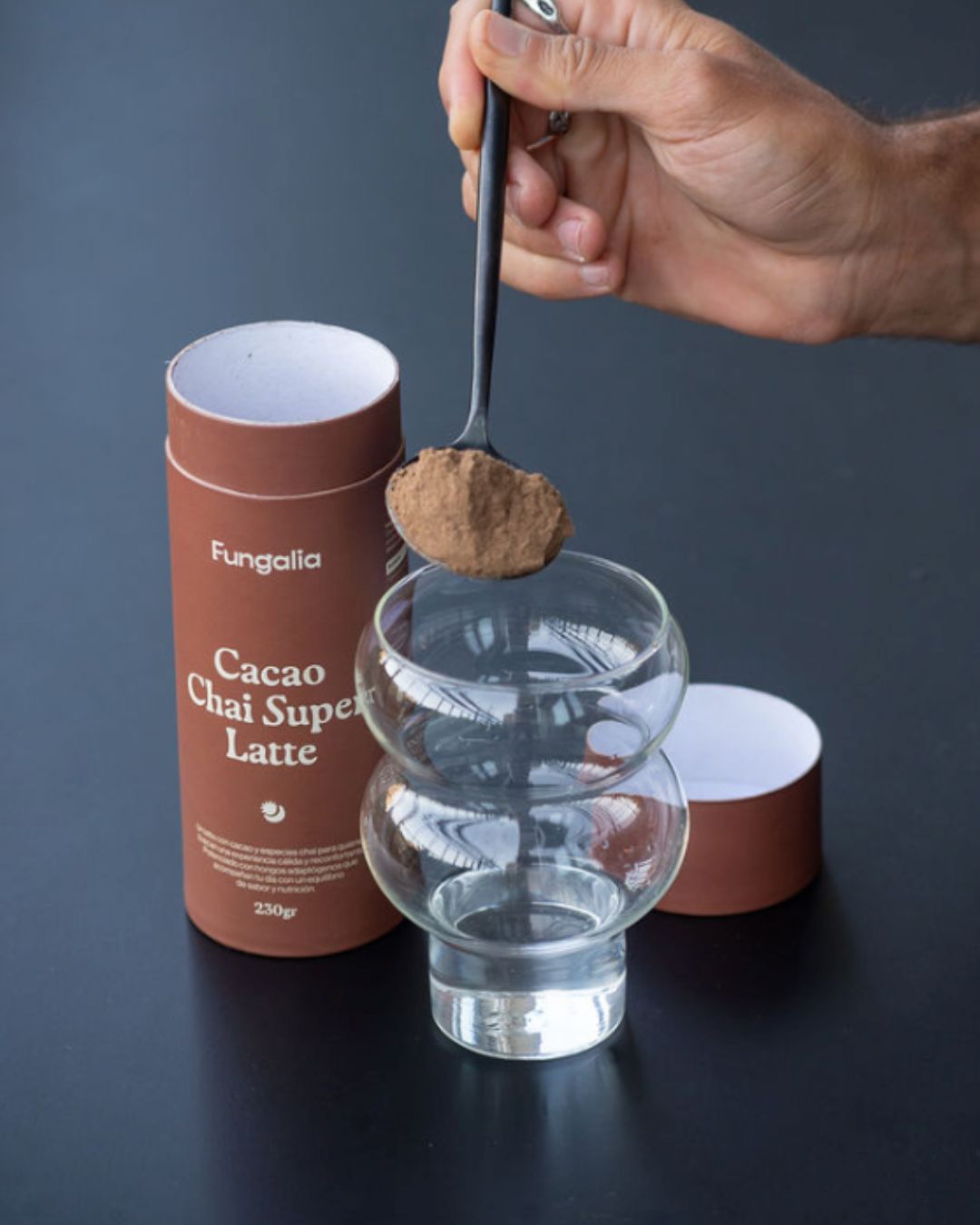 Hand holding spoon with powder over glass, next to Cacao Chai Super Latte container.