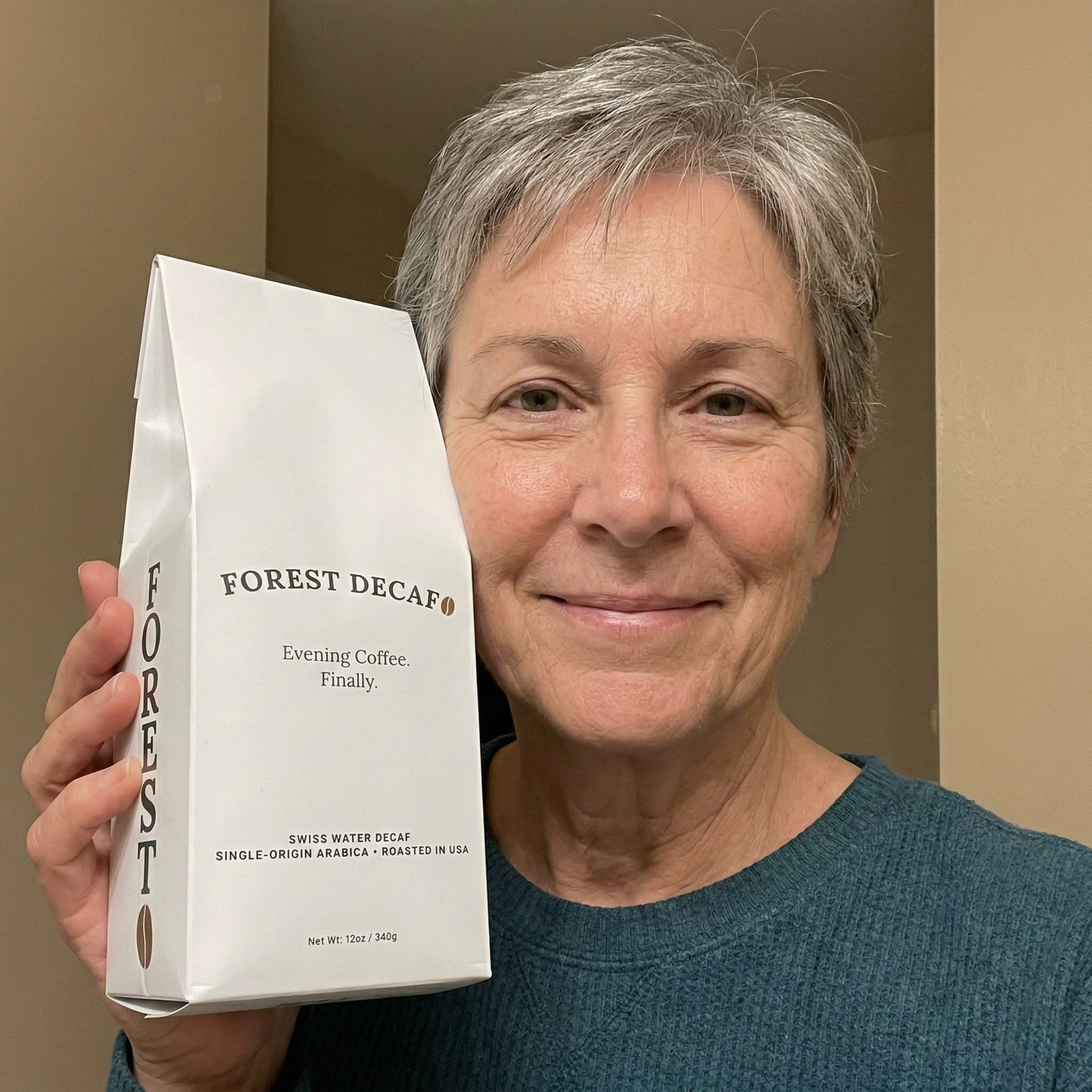 A smiling woman with short gray hair holds up a white bag of Forest Decaf coffee.
