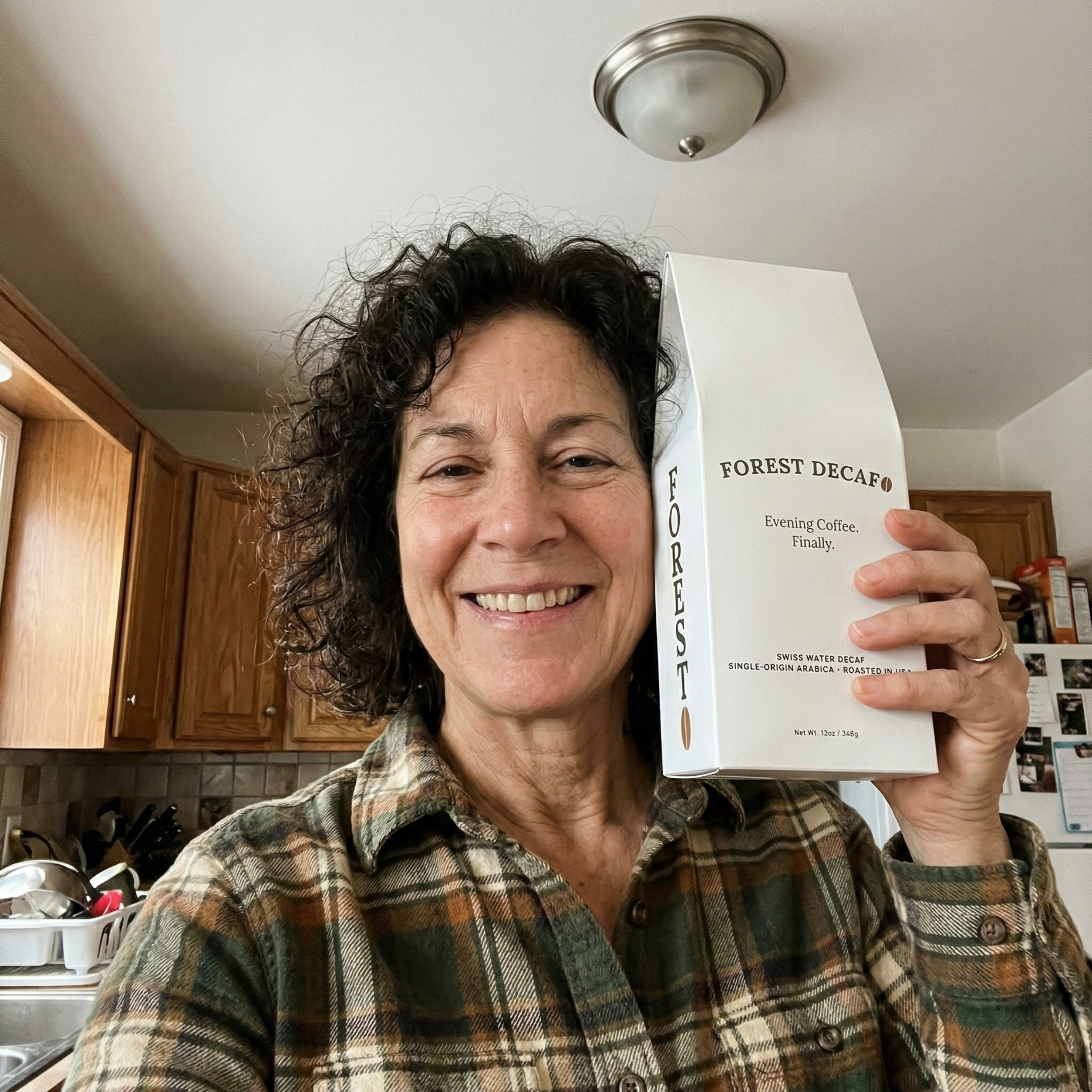 A smiling woman in a plaid shirt holds up a white box of Forest Decaf coffee in a kitchen.