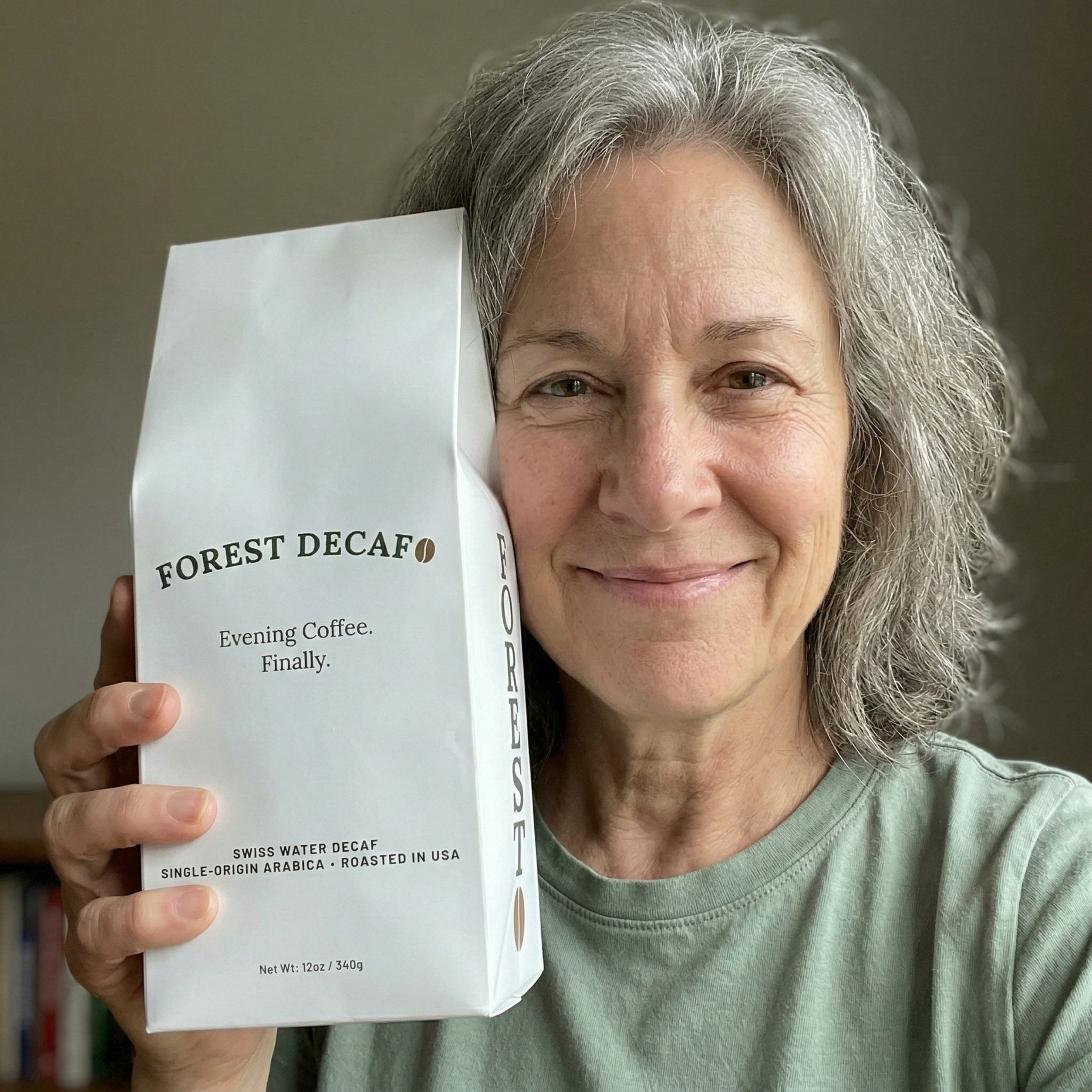 A smiling woman with gray hair holds up a white bag of Forest Decaf coffee.