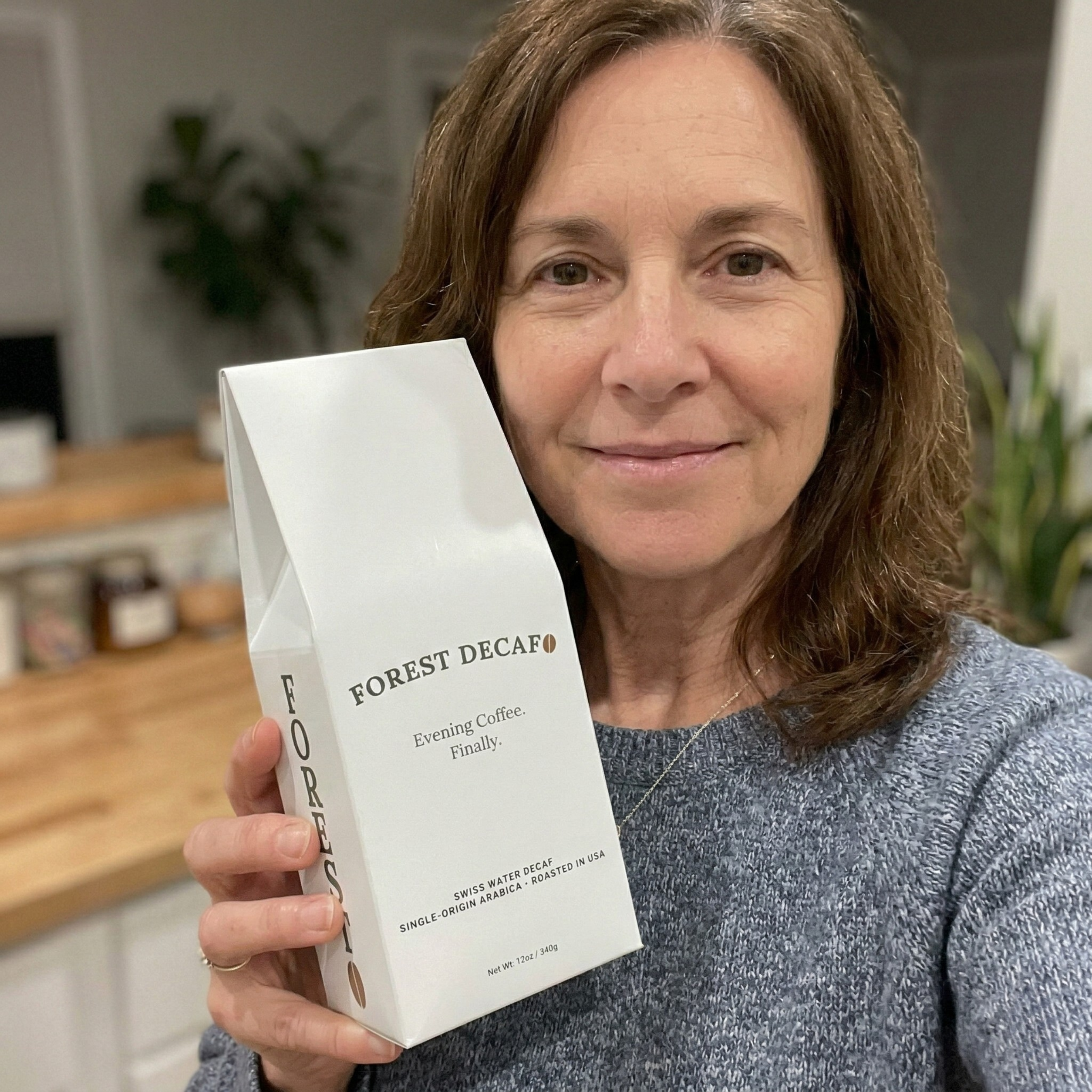 A woman in a grey sweater holds up a white box of Forest Decaf coffee.