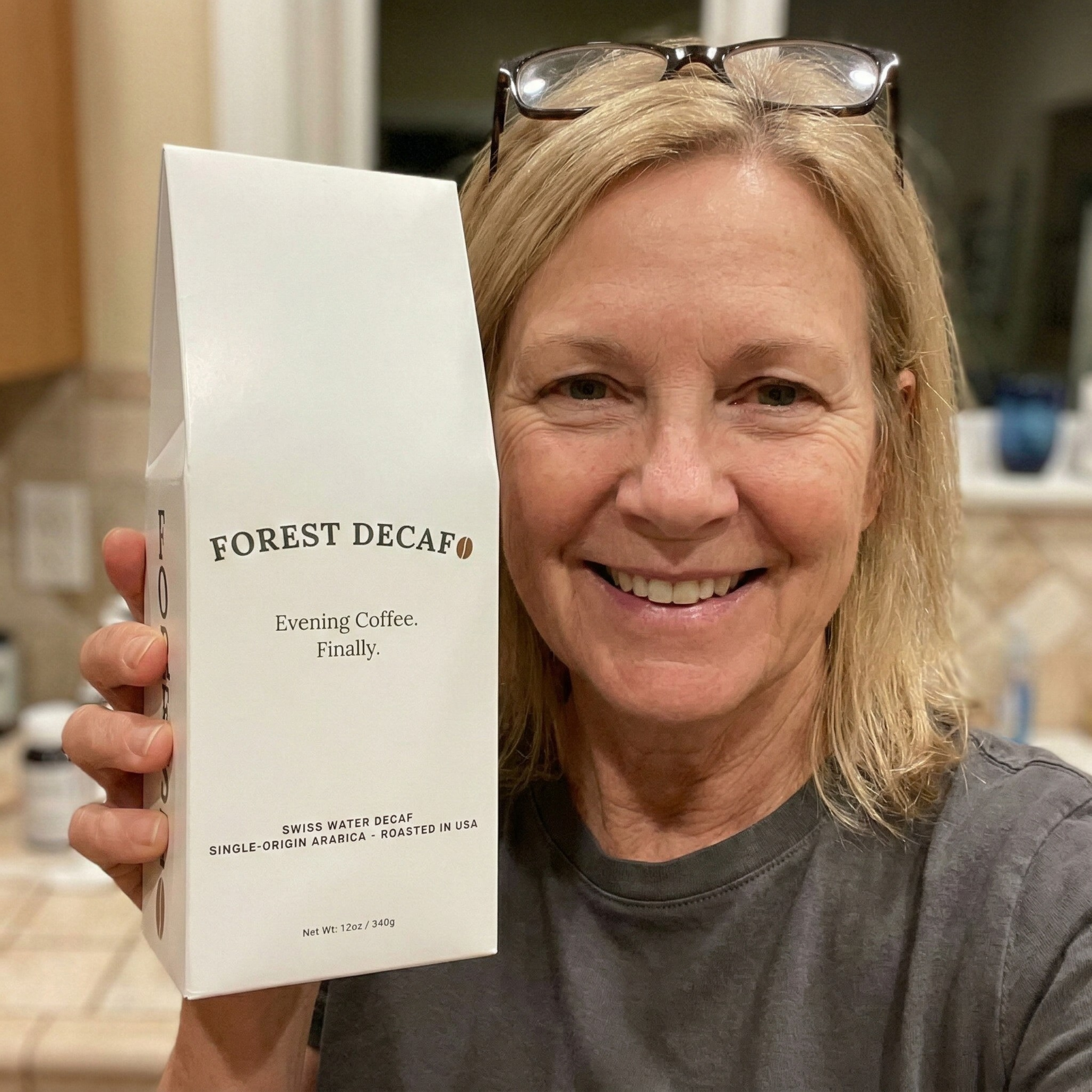 A smiling blonde woman holds up a white box of Forest Decaf coffee in her kitchen.