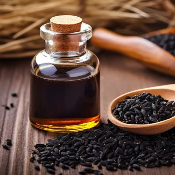 A small glass bottle of dark oil sits on a wooden surface next to black seeds on a wooden spoon.