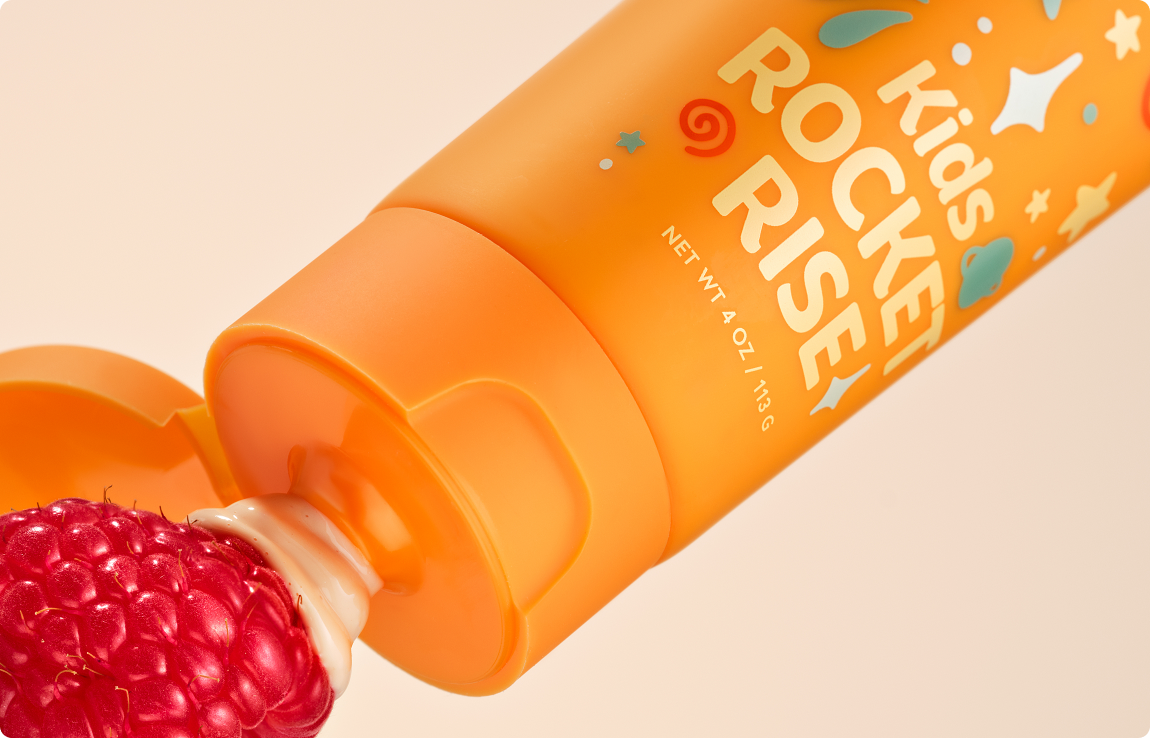 An orange tube of Kids Rocket Rise toothpaste is squeezed onto a fresh raspberry against a peach background.