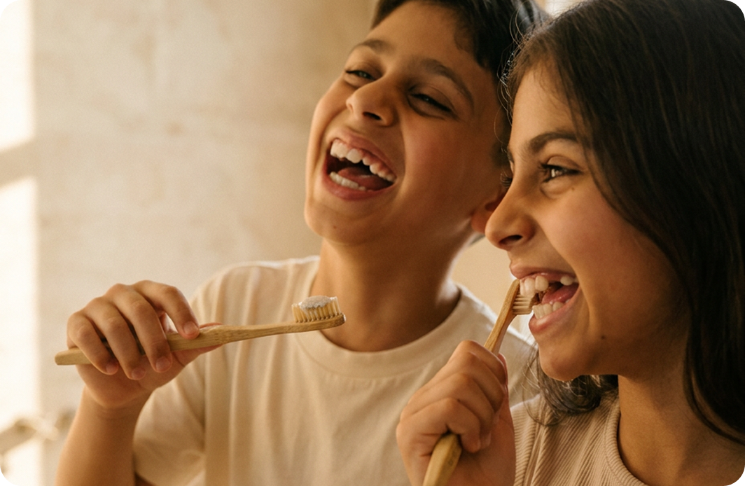 A boy and girl laughing while brushing their teeth with bamboo toothbrushes in a bathroom.