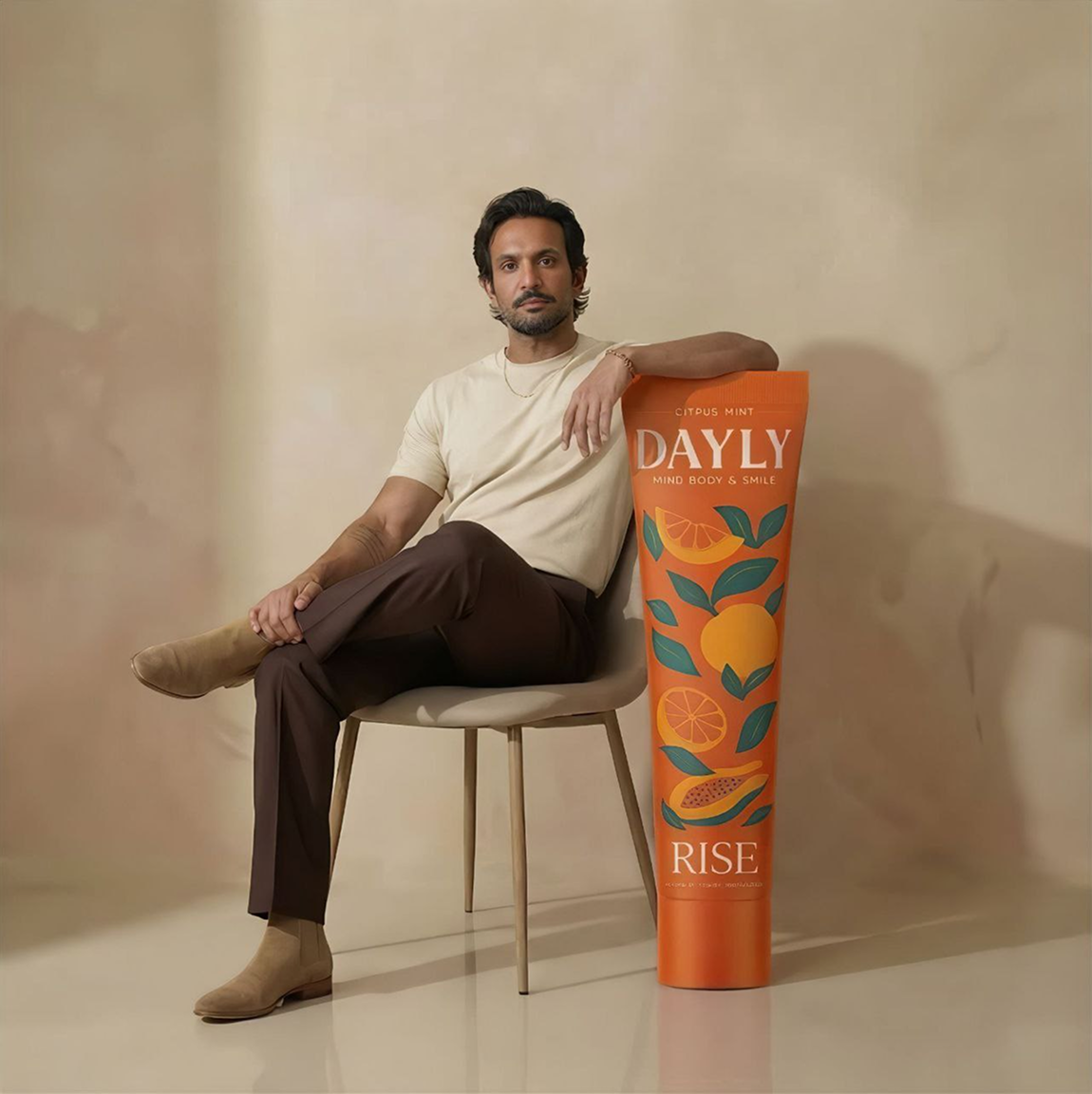 A man sits in a chair, resting his arm on a giant orange tube of Dayly brand lotion.
