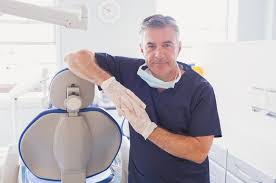 A male dentist in blue scrubs and white gloves leans on a dental chair in a clinic.