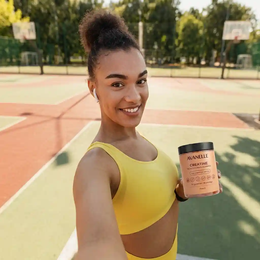 A smiling woman in a yellow sports bra holds a jar of Avanelle creatine on a basketball court.