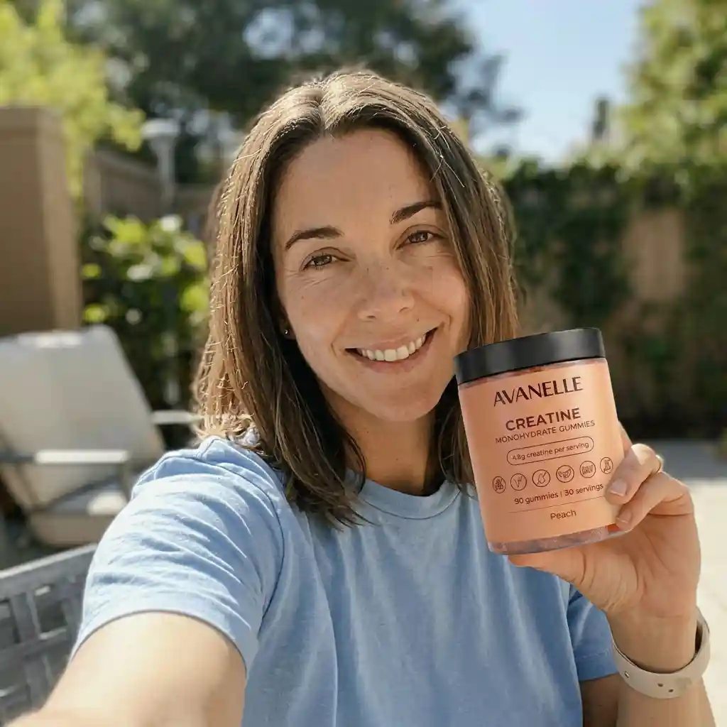 A smiling woman in a blue shirt holds a container of Avanelle Creatine Monohydrate Gummies outdoors.