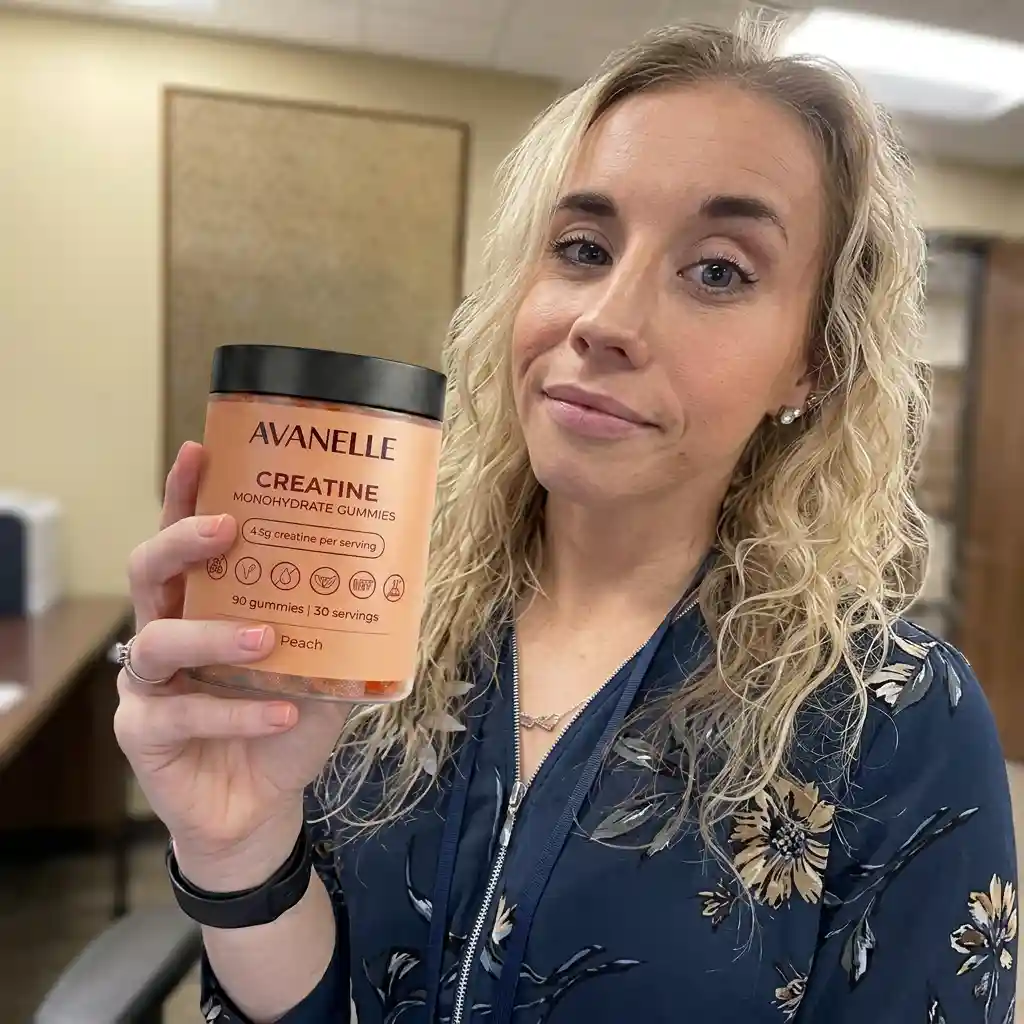 A woman with blonde hair holds up a jar of Avanelle Creatine Monohydrate Gummies in an office setting.