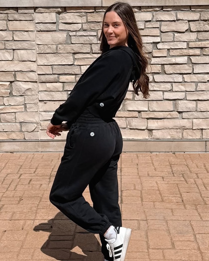 A woman in a black sweatsuit smiles while posing outdoors against a light-colored stone wall.