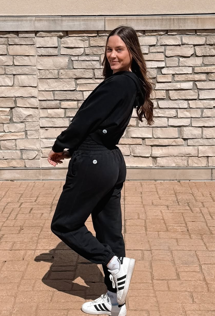 A woman in a black sweatsuit smiles while posing outdoors against a light-colored stone wall.