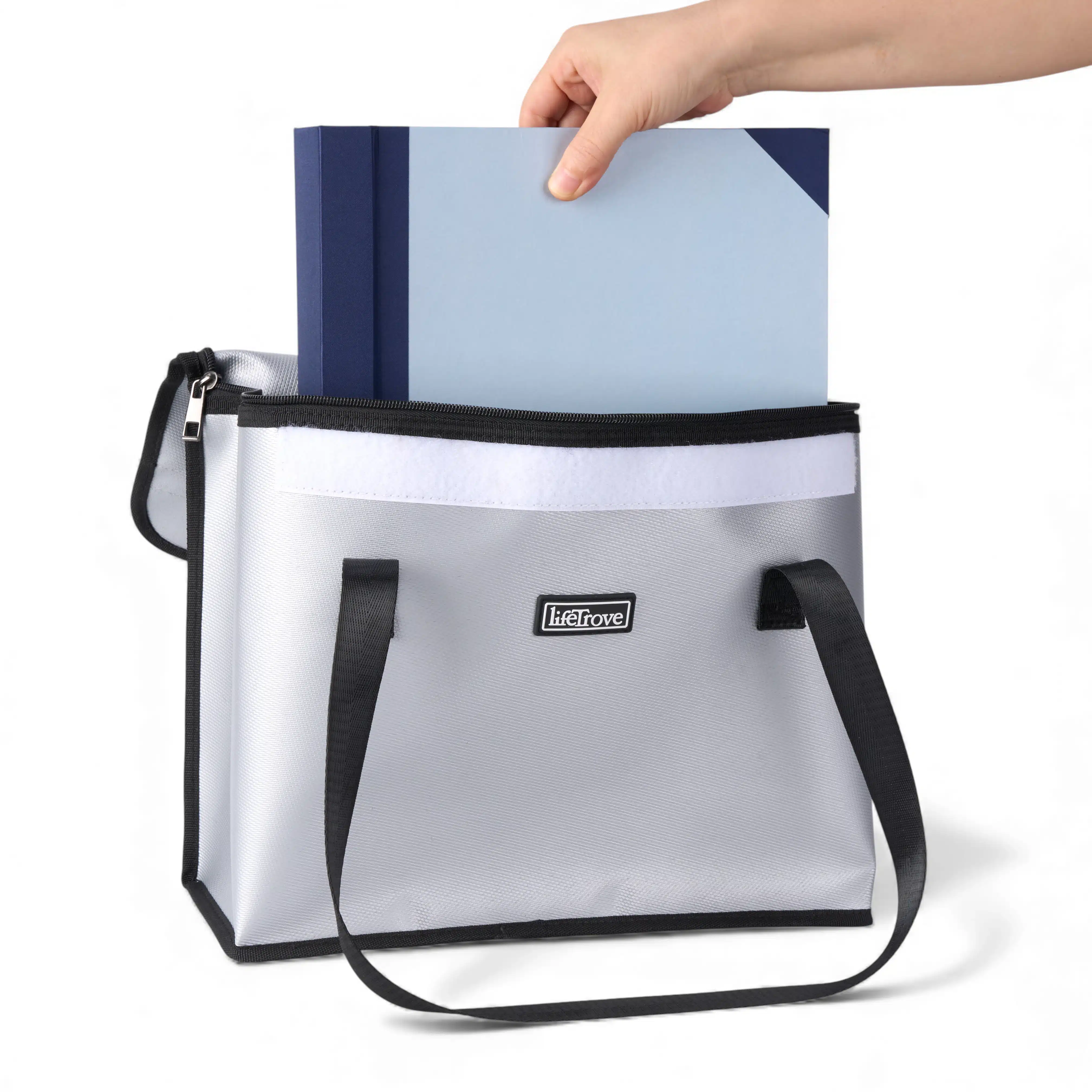 A hand places a blue folder into a silver zippered tote bag with black handles.