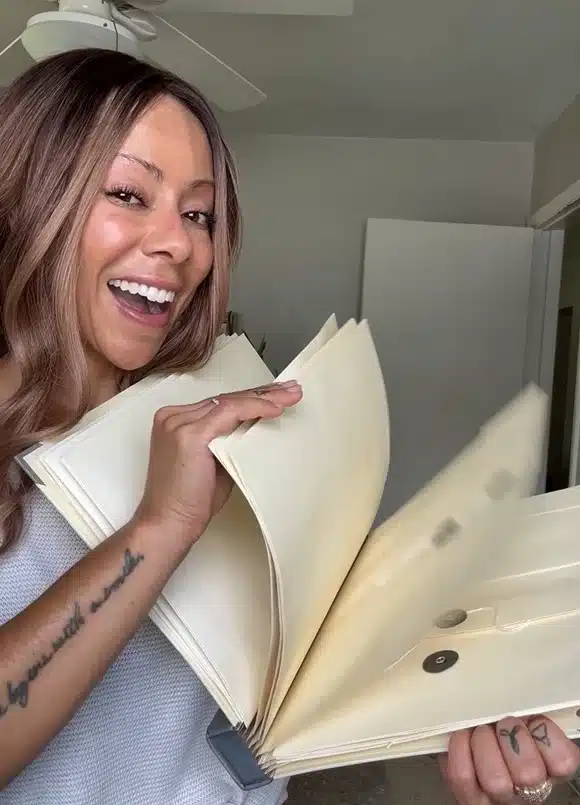 A smiling woman with a tattoo on her arm holds open a large, expanding file folder.