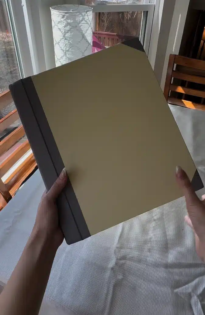 A person's hands holding a large, tan and grey binder or portfolio over a white tablecloth.