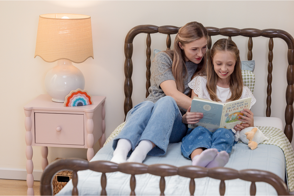 Two people sitting on a bed, reading a book together, with a lamp on a nightstand nearby.