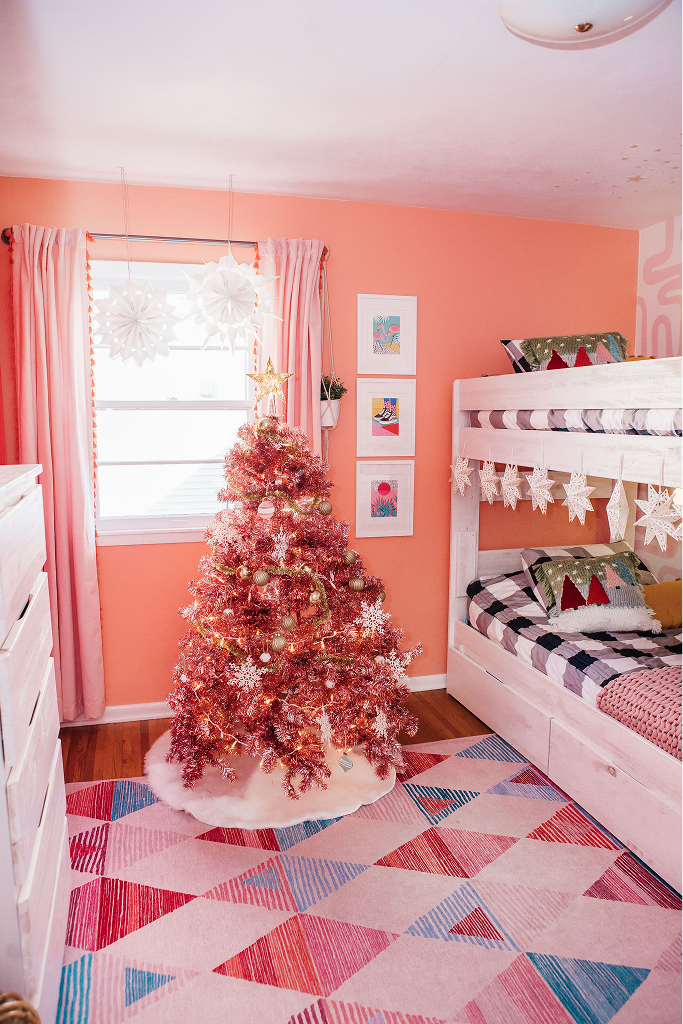 A pink room with white furniture and pink Christmas tree. The tree is decorated with white and gold decor and the room has various snowflake decorations hanging from the ceiling and furniture.