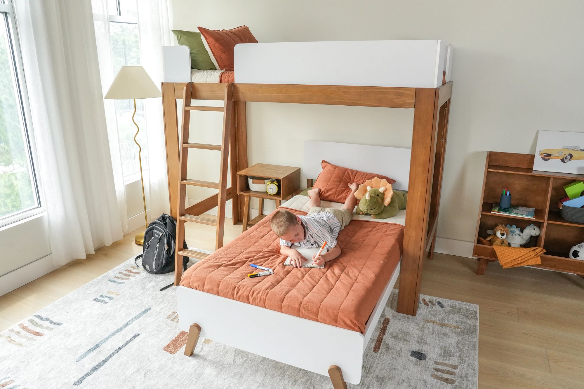 A child laying on their stomach on the bottom bunk of a modern-style, L-shaped bunk bed sketching in a notebook.