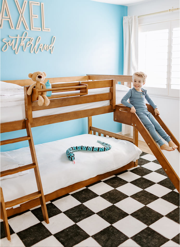 Child sitting on a wooden bunk bed with a slide in a room with a blue wall and checkered floor.