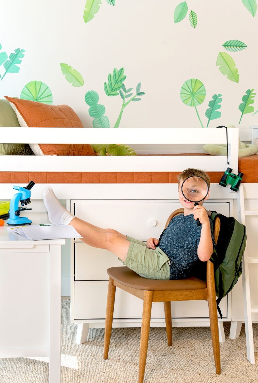 Little boy holding a magnifying glass by his white bunk bed in his jungle explorer themed room with plant watercolor wall decals.