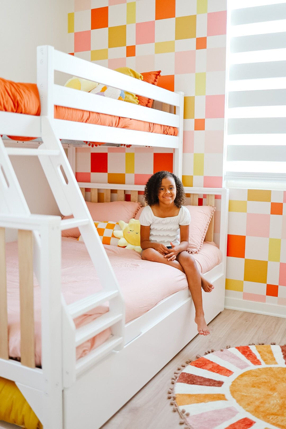 A young girl sitting cross-legged on the bottom bunk of a white bunk bed against a vibrant geometric patterned wall featuring squares in orange, yellow, pink, and coral colors. She is wearing a white top and smiling at the camera.