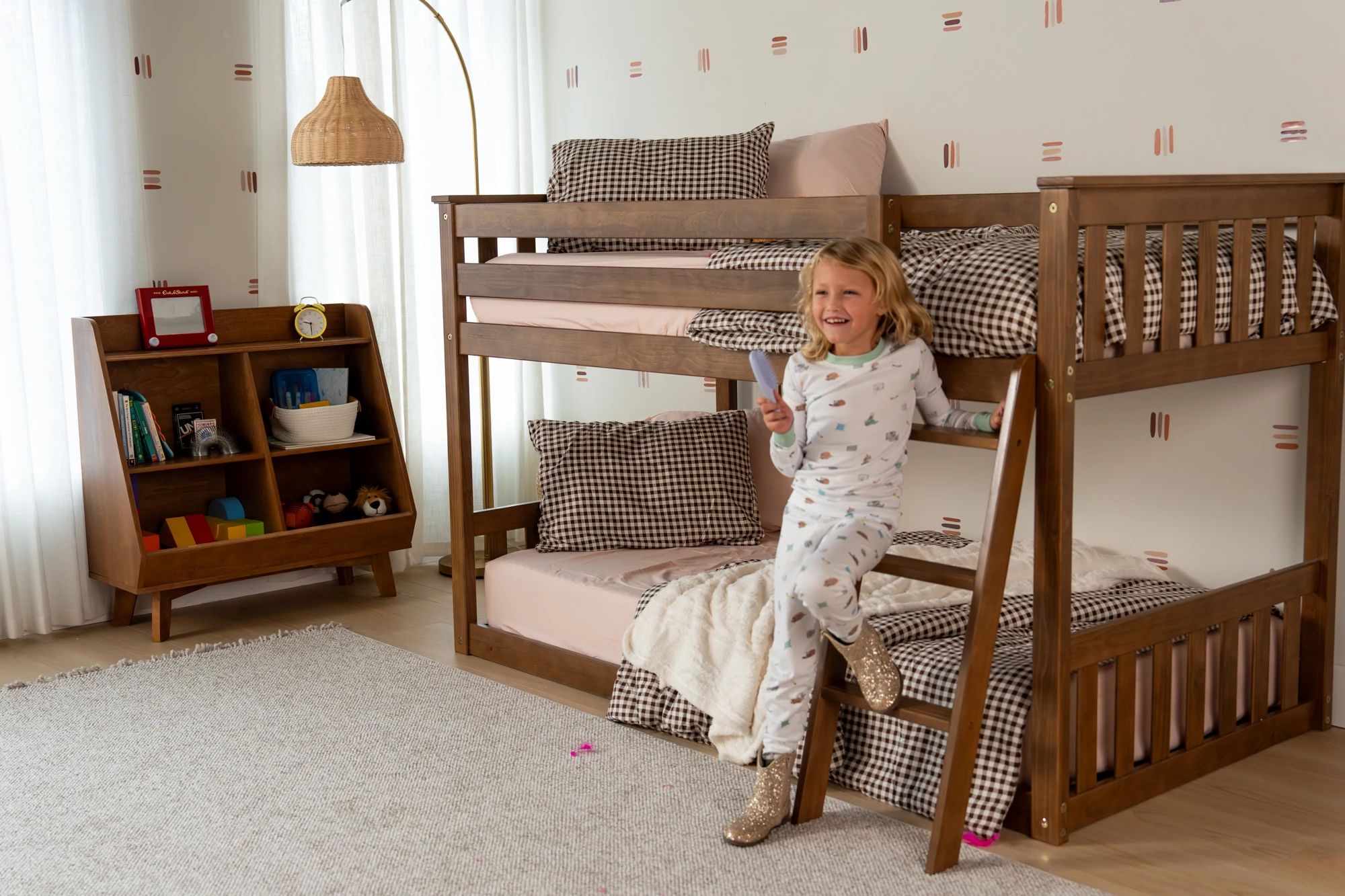 Young child climbing down from a short bunk bed with a pink and brown bedding in a neutral colored bedroom with a bookcase, lamp, and rug.