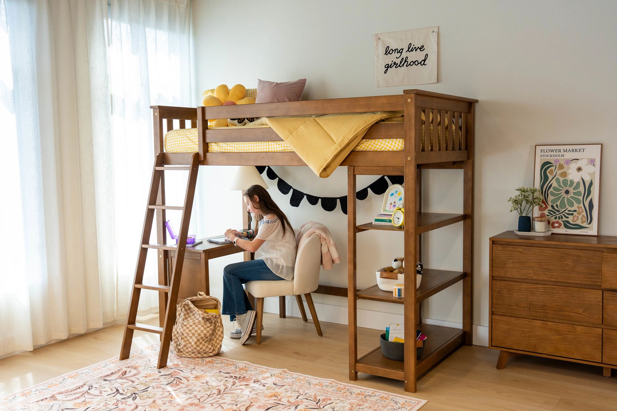 A teen sitting at a desk under a wooden loft bed decorated with yellow and blue decor in a bright room.