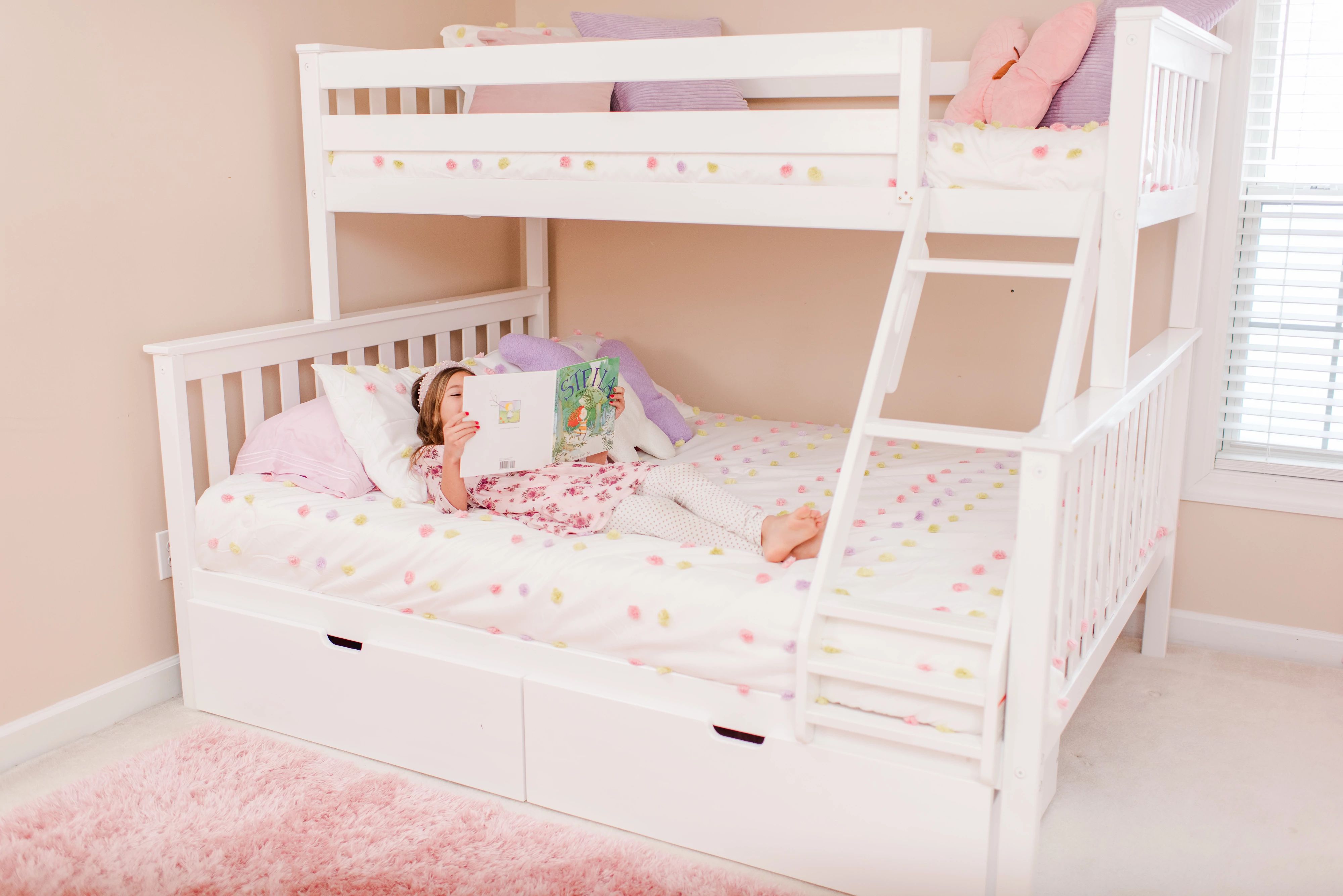 Young child reading a book on the bottom bunk of a white bunk bed in a light pink decorated room.