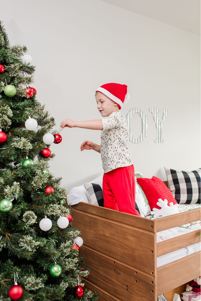 A young boy in red and white holiday attire standing on a low loft bed, placing an ornament on a tree.