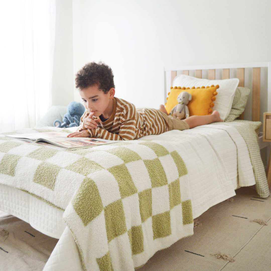 Child reading a book on a bed with checkered blanket and stuffed toys.