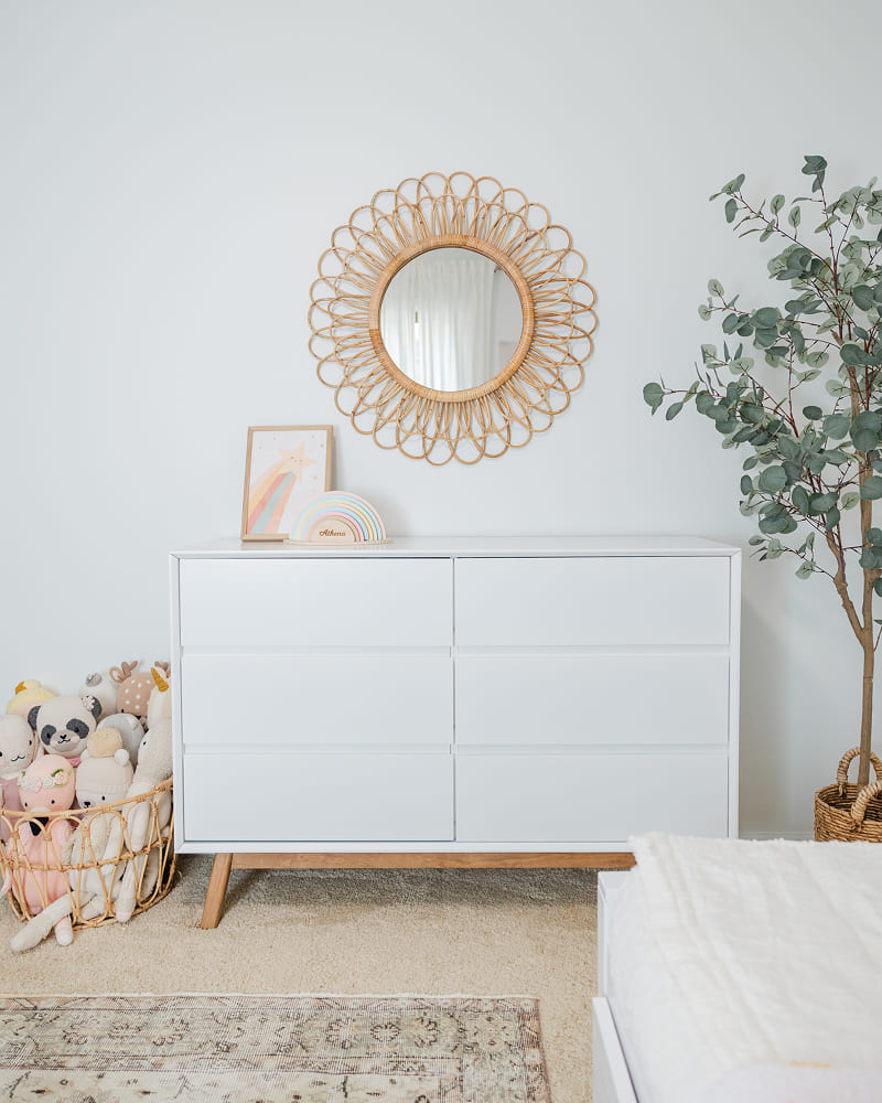 A white 6-drawer dresser in a white room. There is a decorative mirror on the wall above the dresser, and basket of stuffed toys next to it. There is also some pastel rainbow decor on the dresser.