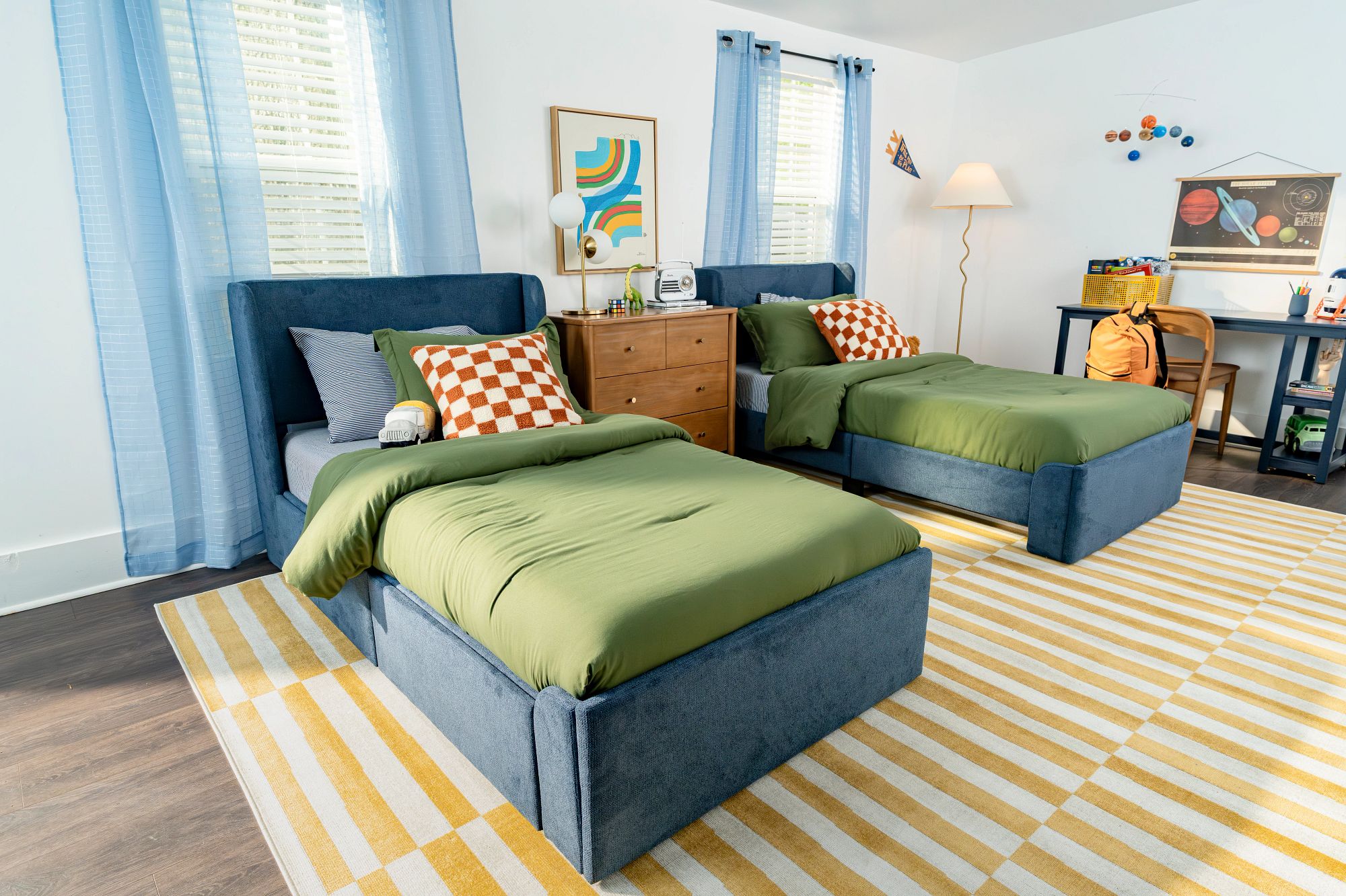 Two blue upholstered twin beds with green and blue bedding in room with a yellow rug, matching blue desk, and space-themed decor.