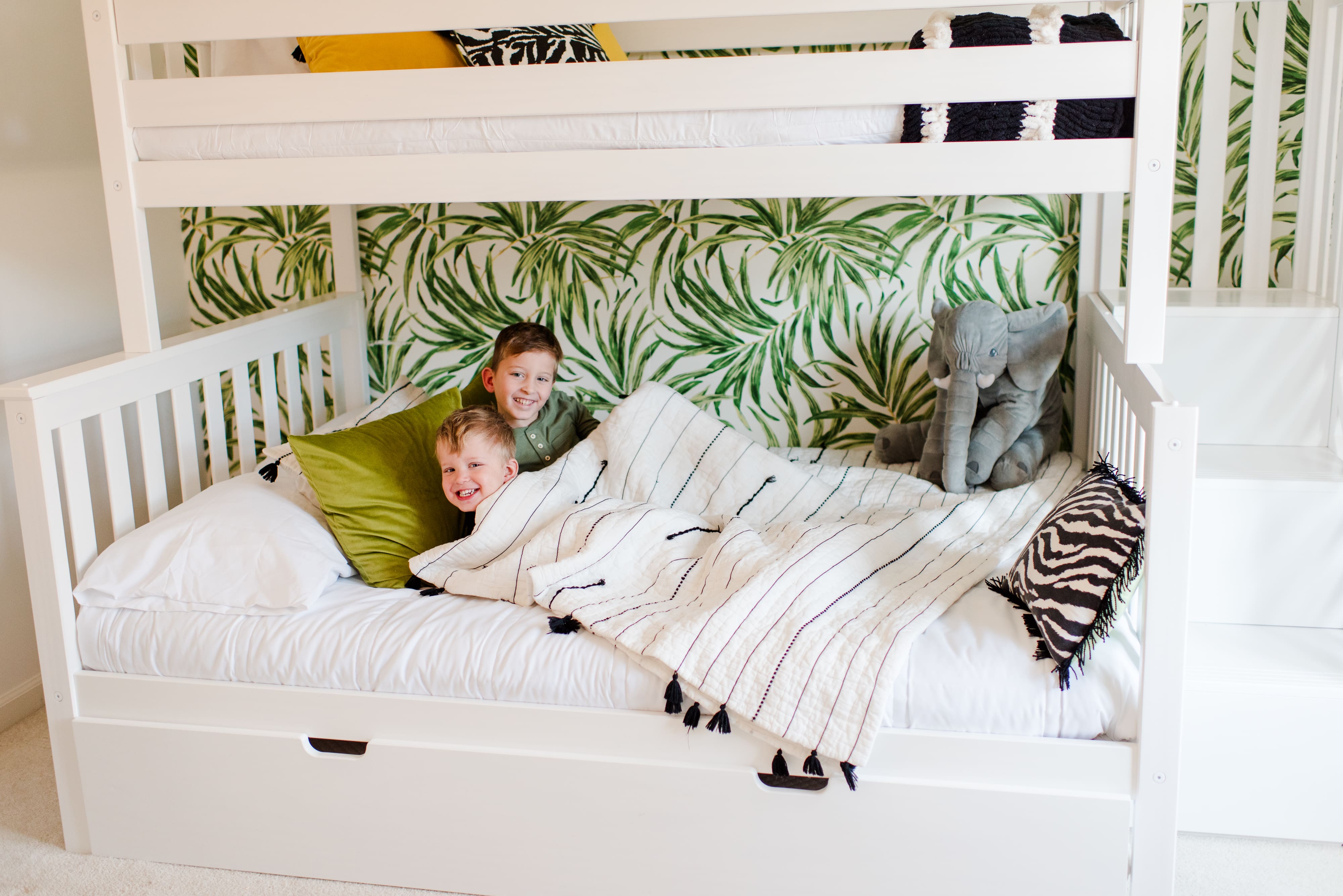 Two children lying in a white bunk bed with tropical palm leaf wallpaper. The older child sits behind the younger child under a white and black striped throw blanket with tassels.