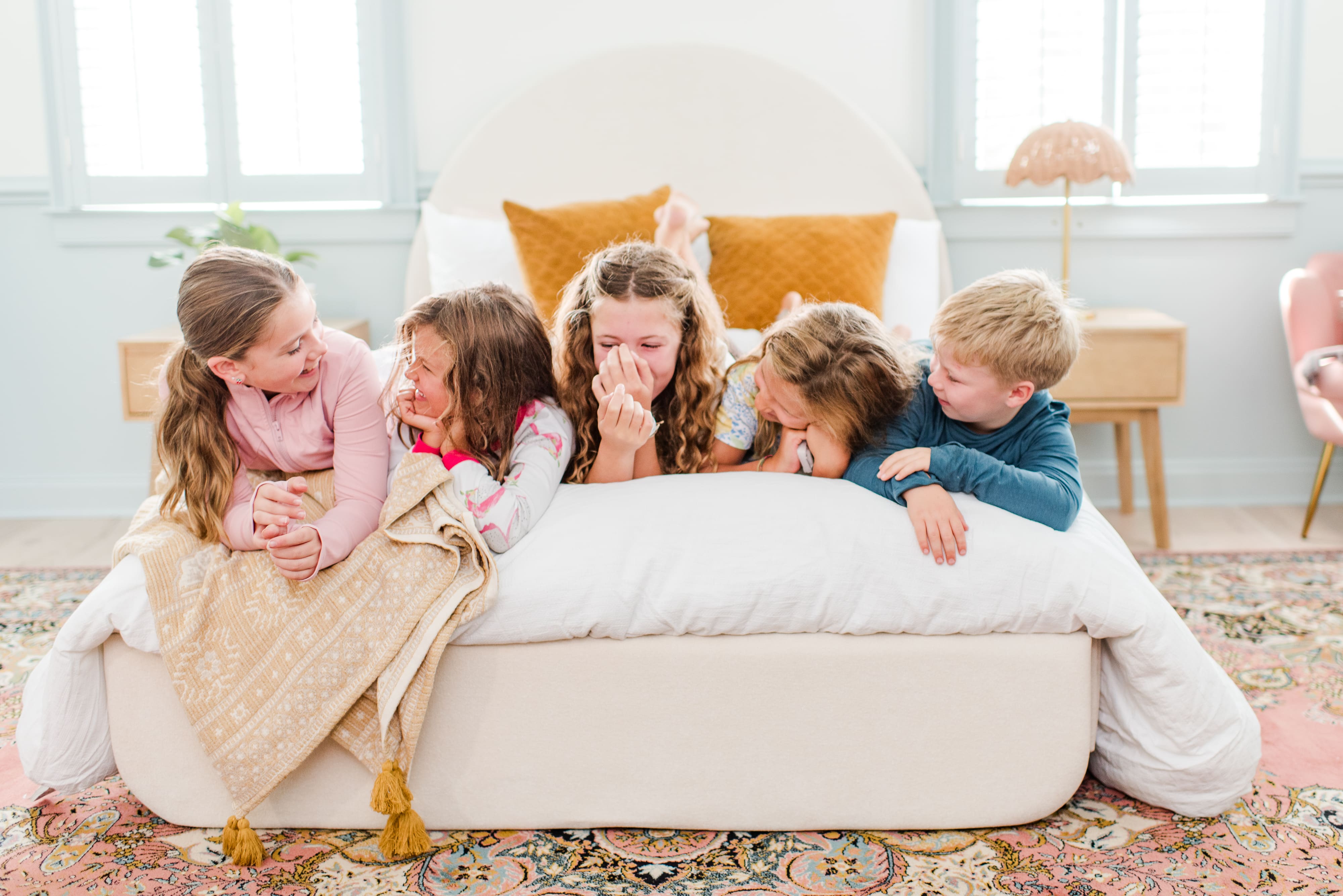 Five children gathered around a large white floor cushion or bean bag in a bright living room. They are all leaning on or hugging the cushion, with one child covering their face with their hands. The room features a white sofa with orange pillows and large windows in the background.