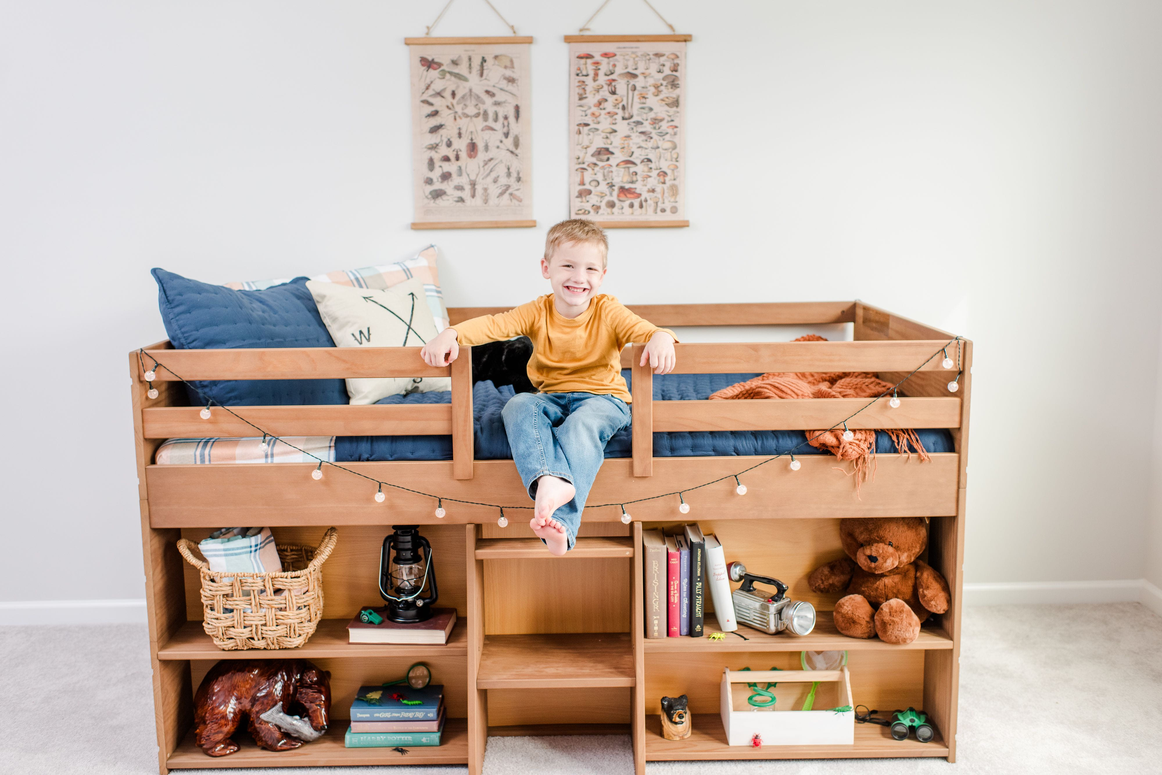 A blonde boy in an orange shirt and jeans sitting on a wooden loft bed that features built-in storage shelves underneath. The shelves contain books, baskets, toys including a teddy bear, and other items. String lights are draped around the bed frame, and educational posters hang on the white wall behind.