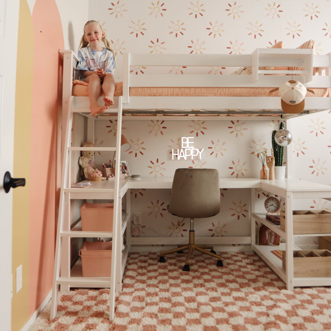 Child sitting on a loft bed in a decorated room with a desk underneath.