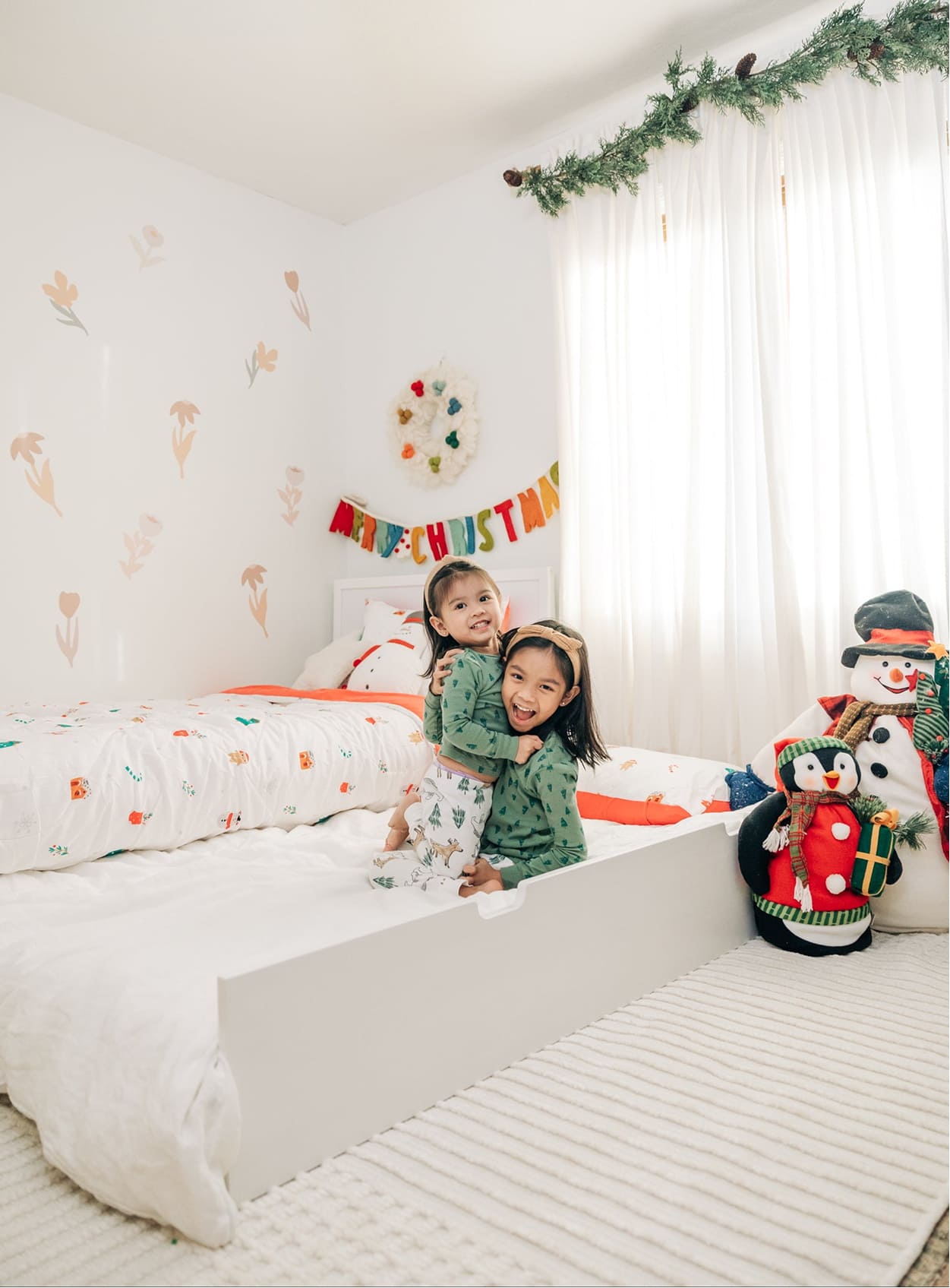 Two smiling children in matching green Christmas pajamas hugging on a white toddler bed in a bright, holiday-decorated kids’ bedroom with a “Merry Christmas” banner, garland, plush penguin and snowman toys, white curtains and floral wall decals