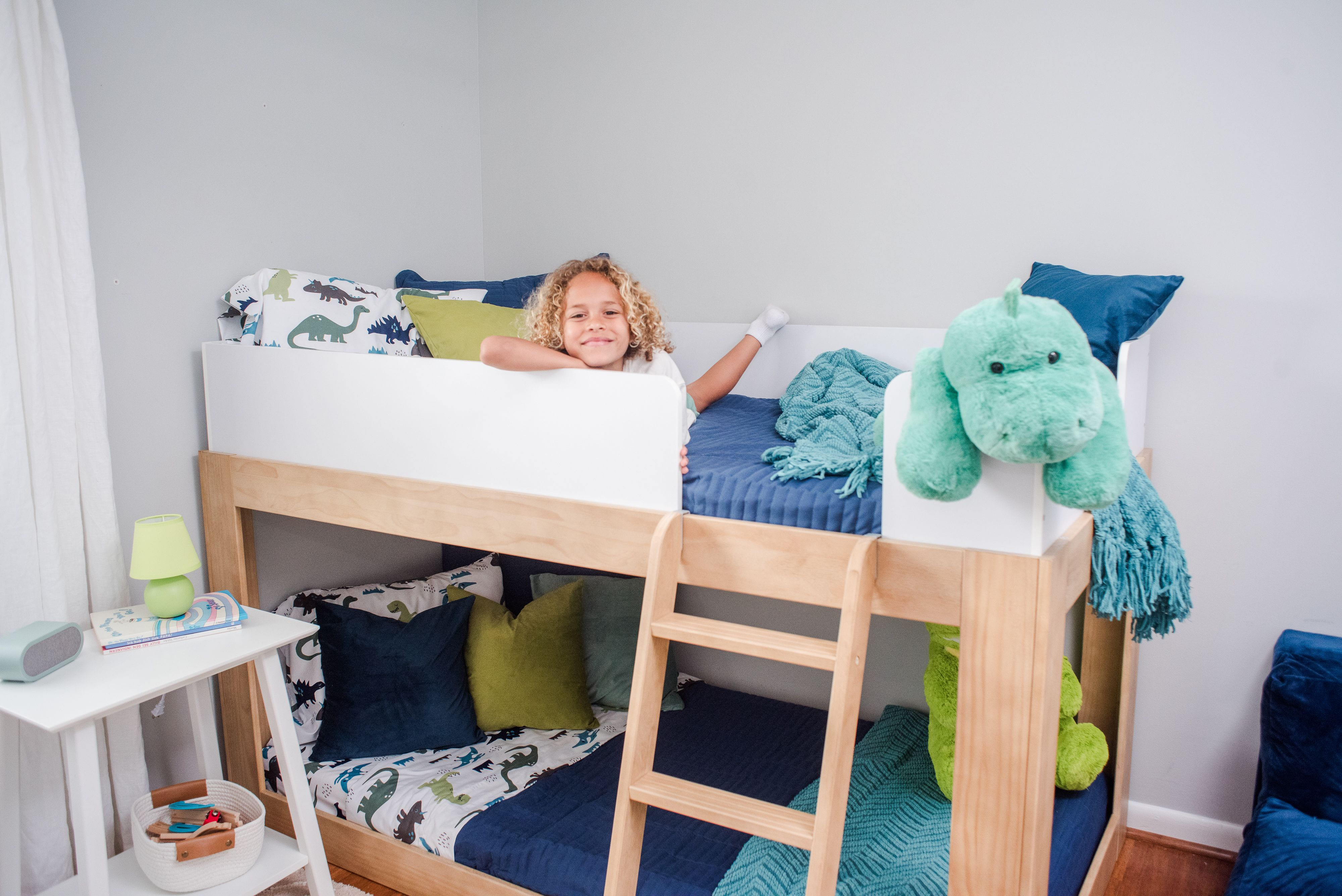 A curly-haired child lying on the top bunk of a wooden bunk bed, holding a book and smiling. The bed is decorated with blue bedding, dinosaur-themed pillows, and a large green dinosaur stuffed animal.